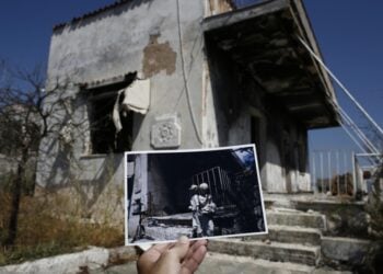 Φωτορεπόρτερ κρατάει φωτογραφία, που τραβήχτηκε ένα χρόνο μετά τη θανατηφόρα πυρκαγιά της 23ης Ιουλίου 2018 στο Μάτι και δείχνει αγαλματίδια σε καμένη αυλή του ίδιου σπιτιού που απεικονίζεται στη μεγαλύτερη φωτογραφία (φωτ.: ΑΠΕ-ΜΠΕ/Γιάννης Κολεσίδης)