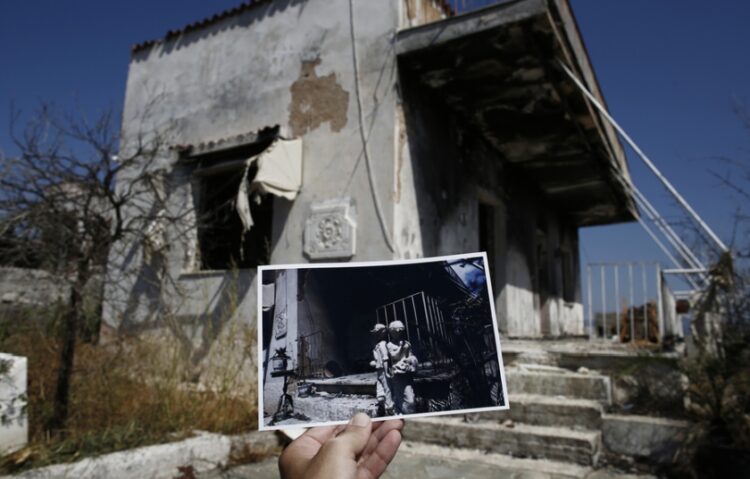 Φωτορεπόρτερ κρατάει φωτογραφία, που τραβήχτηκε ένα χρόνο μετά τη θανατηφόρα πυρκαγιά της 23ης Ιουλίου 2018 στο Μάτι και δείχνει αγαλματίδια σε καμένη αυλή του ίδιου σπιτιού που απεικονίζεται στη μεγαλύτερη φωτογραφία (φωτ.: ΑΠΕ-ΜΠΕ/Γιάννης Κολεσίδης)