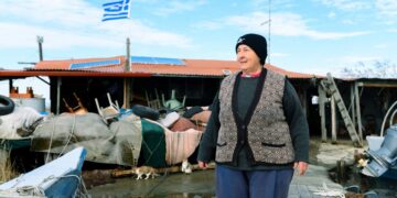 Η Άρτεμις Παπακωστίδου, γνωστή ως «Κυρά του Δέλτα», μπροστά από την «καλύβα» που διατηρεί με τον σύζυγό της, με την ελληνική σημαία να ανεμίζει στο βάθος (φωτ.: ΑΠΕ-ΜΠΕ/Δημήτρης Αλεξούδης)