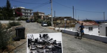 Ο φωτορεπόρτερ κρατάει φωτογραφία, που τραβήχτηκε μια ημέρα μετά την πυρκαγιά και δείχνει μια γυναίκα να περπατάει μπροστά από καμένα αυτοκίνητα, στο Μάτι, ενώ ένα αγόρι περπατάει με το ποδήλατό του στο ίδιο σημείο, στο παραθαλάσσιο θέρετρο, έναν χρόνο μετά (φωτ.: ΑΠΕ-ΜΠΕ/Γιάννης Κολεσίδης)