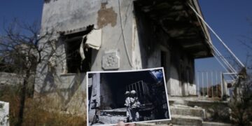 Φωτορεπόρτερ κρατάει φωτογραφία, που τραβήχτηκε ένα χρόνο μετά τη θανατηφόρα πυρκαγιά της 23ης Ιουλίου 2018 στο Μάτι και δείχνει αγαλματίδια σε καμένη αυλή του ίδιου σπιτιού που απεικονίζεται στη μεγαλύτερη φωτογραφία (φωτ.: ΑΠΕ-ΜΠΕ/Γιάννης Κολεσίδης)