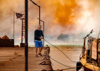 Άνδρας δένει βάρκα στο λιμάνι Λιθίου στη Χίο, ενώ πυρκαγιά καίει δασική έκταση στο βάθος (φωτ.: ΑΠΕ-ΜΠΕ/Κώστας Κούργιας)