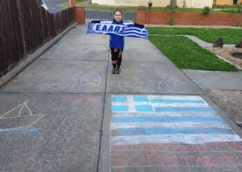 Μικρούλα ομογενής και μαθήτρια ελληνικού σχολείου στη Μελβούρνη κρατά κασκόλ με την ελληνική σημαία, μπροστά από ζωγραφιές με ελληνικό θέμα στο πλακόστρωτο (φωτ.: facebook/Modern Greek Teachers' Association of Victoria)