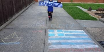 Μικρούλα ομογενής και μαθήτρια ελληνικού σχολείου στη Μελβούρνη κρατά κασκόλ με την ελληνική σημαία, μπροστά από ζωγραφιές με ελληνικό θέμα στο πλακόστρωτο (φωτ.: facebook/Modern Greek Teachers' Association of Victoria)