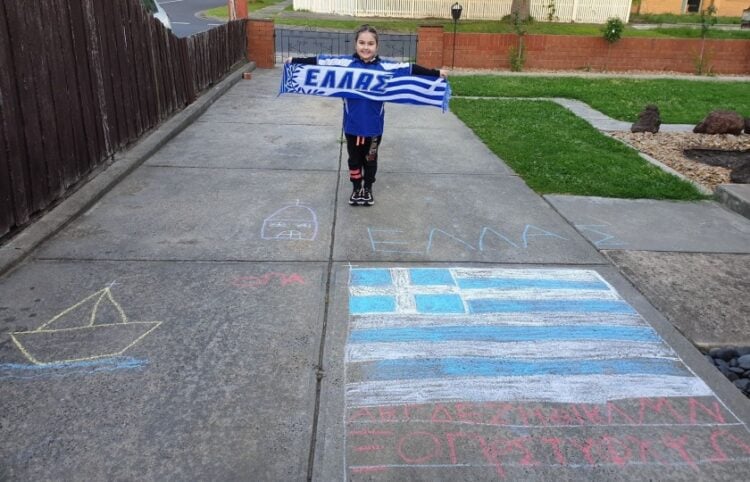 Μικρούλα ομογενής και μαθήτρια ελληνικού σχολείου στη Μελβούρνη κρατά κασκόλ με την ελληνική σημαία, μπροστά από ζωγραφιές με ελληνικό θέμα στο πλακόστρωτο (φωτ.: facebook/Modern Greek Teachers' Association of Victoria)