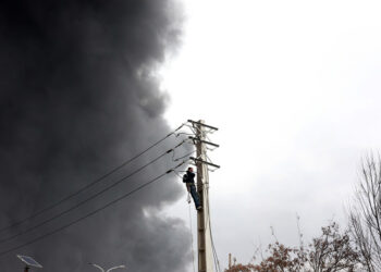 Ιρανός εργάτης επισκευάζει δίκτυο ηλεκτροδότησης, ενώ καπνός εξακολουθεί να υψώνεται από το διυλιστήριο πετρελαίου Shahran μετά από αεροπορική επιδρομή στην Τεχεράνη. 8 Μαρτίου 2026 (φωτ.: EPA / Abedin Taherkenareh)