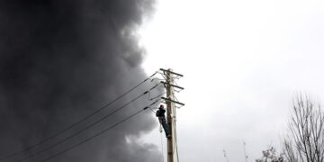 Ιρανός εργάτης επισκευάζει δίκτυο ηλεκτροδότησης, ενώ καπνός εξακολουθεί να υψώνεται από το διυλιστήριο πετρελαίου Shahran μετά από αεροπορική επιδρομή στην Τεχεράνη. 8 Μαρτίου 2026 (φωτ.: EPA / Abedin Taherkenareh)