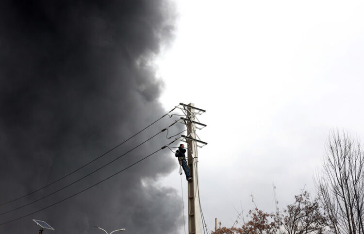 Ιρανός εργάτης επισκευάζει δίκτυο ηλεκτροδότησης, ενώ καπνός εξακολουθεί να υψώνεται από το διυλιστήριο πετρελαίου Shahran μετά από αεροπορική επιδρομή στην Τεχεράνη. 8 Μαρτίου 2026 (φωτ.: EPA / Abedin Taherkenareh)